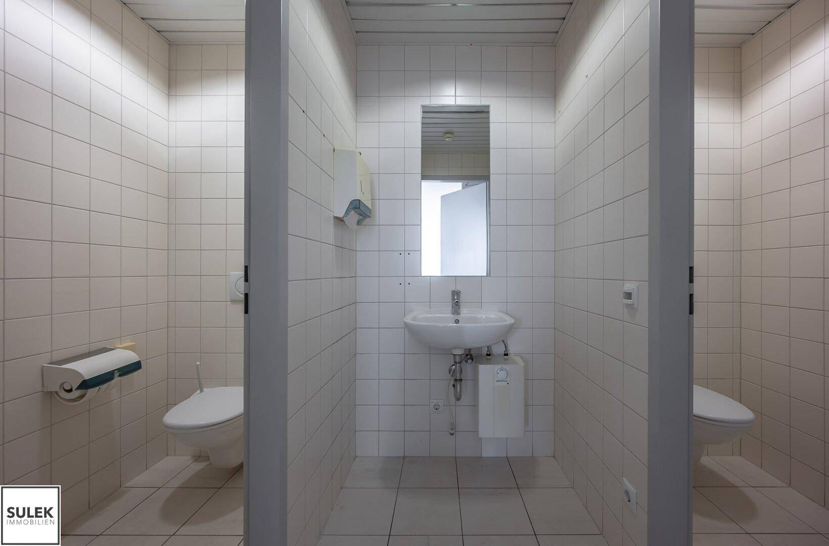 Gefliester Toilettenraum mit Waschbecken und zwei separaten Kabinen.