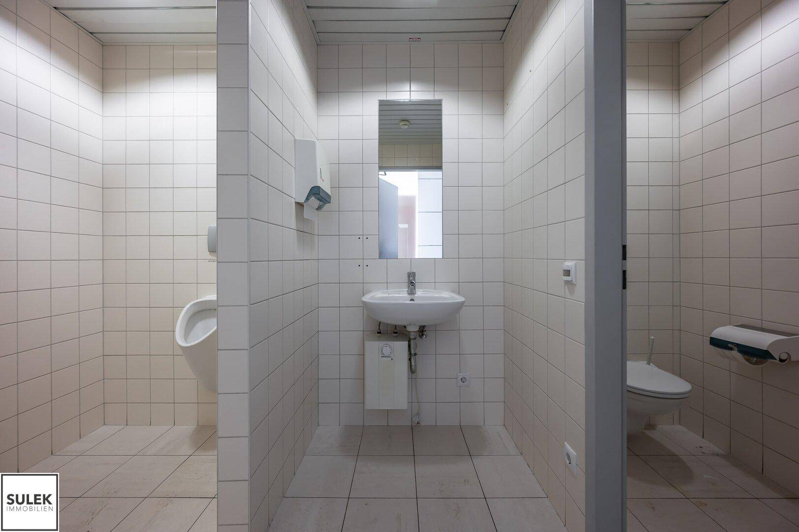 Gefliester Toilettenraum mit Waschbecken, Urinal und separaten Kabinen.