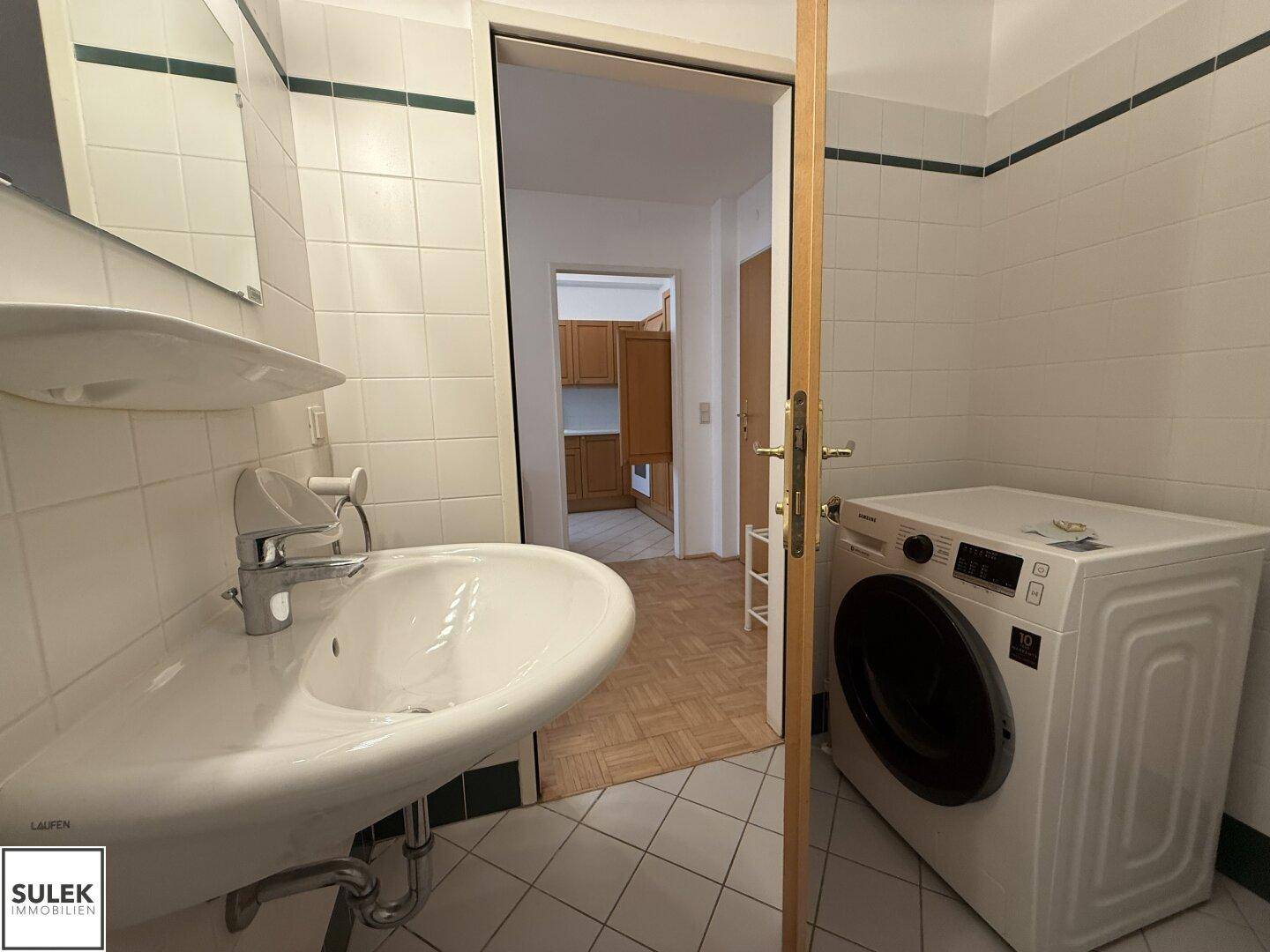 Badezimmer mit Waschbecken, Waschmaschine und Fliesenboden, Blick zur Küche.