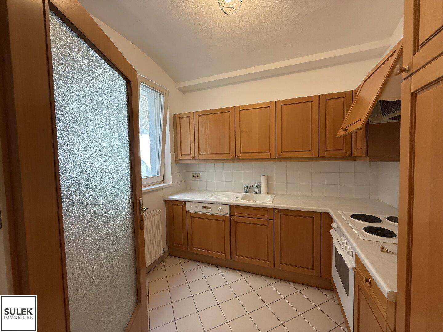 Helle Küche mit Holzschränken, Spüle unter dem Fenster und Fliesenboden.
