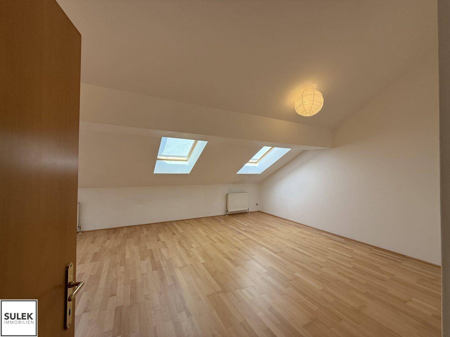 Helles Dachzimmer mit schrägen Wänden, Holzboden und zwei Dachfenstern für viel Tageslicht.