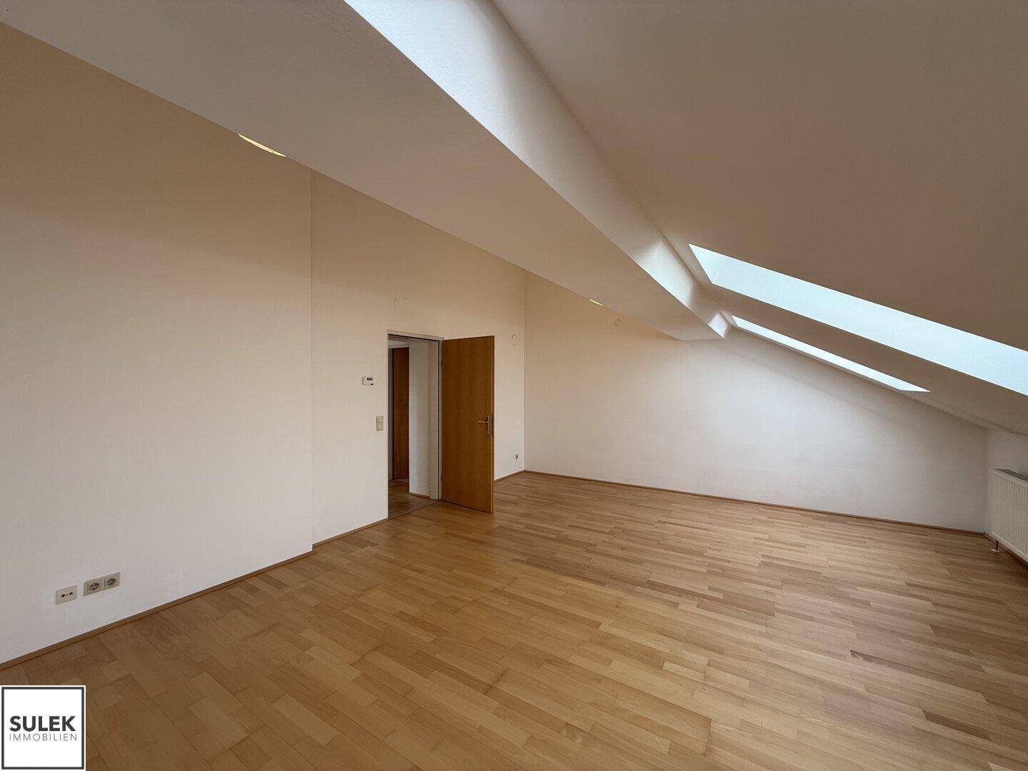Geräumiges Dachzimmer mit Holzboden, schrägen Wänden und mehreren Dachfenstern.