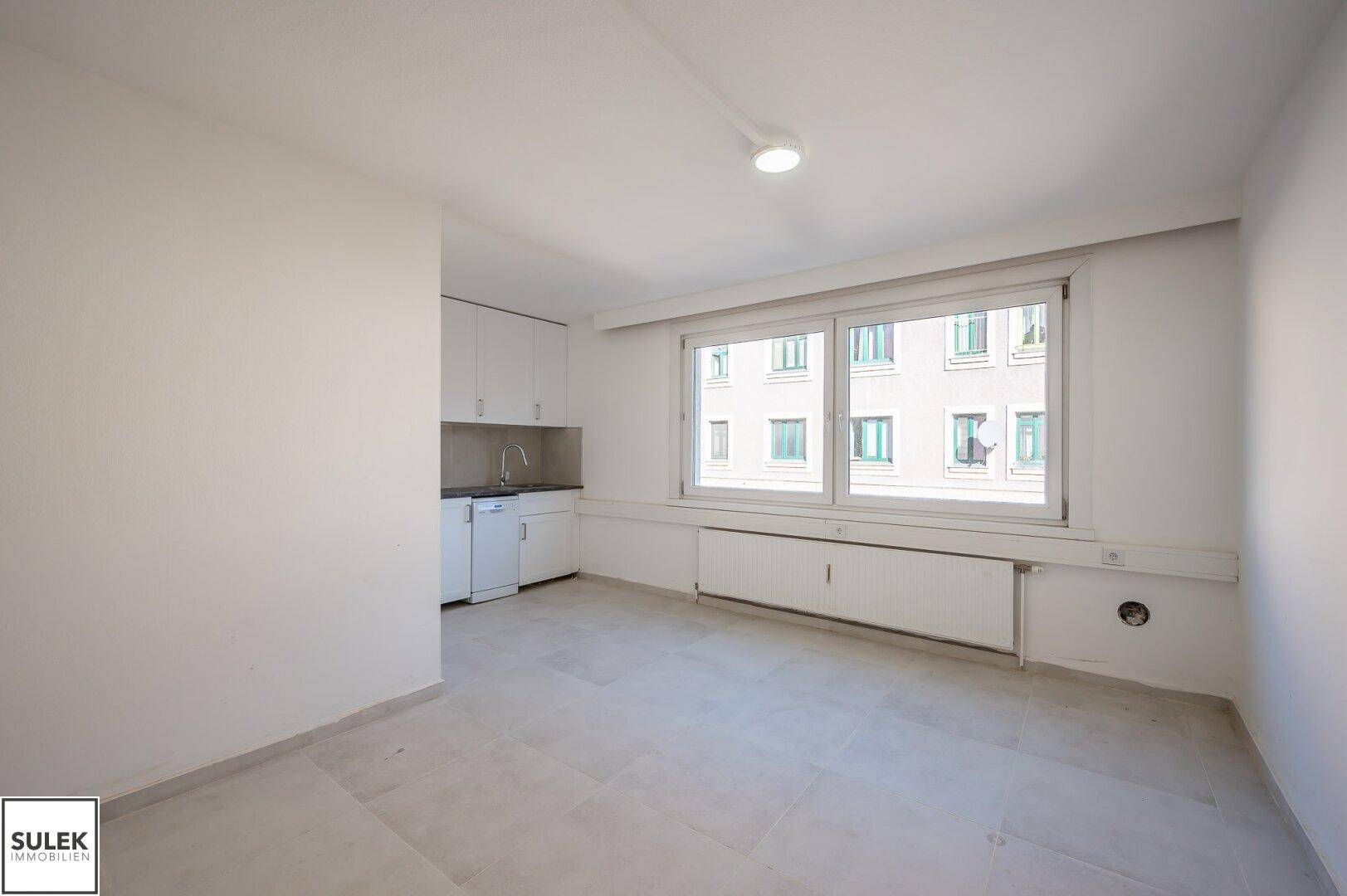 Kleiner Küchenbereich mit weißen Schränken, Spüle und Fliesenboden in einem hellen Raum.