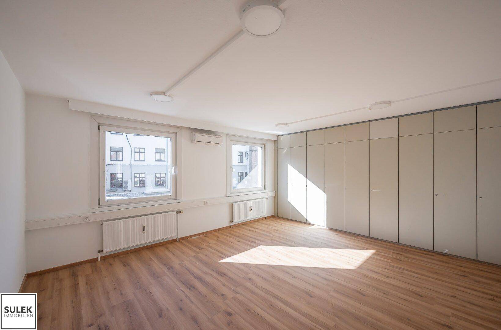 Heller Büroraum mit Laminatboden, Einbauschränken und Fenstern mit Blick auf die Umgebung.