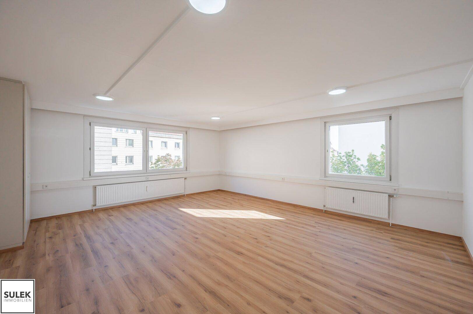 Heller Büroraum mit Laminatboden und großen Fenstern, die viel Tageslicht hereinlassen.