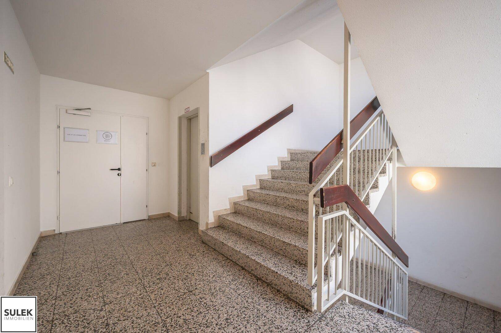 Treppenhaus mit Steinstufen, Metallgeländer und Aufzugszugang, gut beleuchtet.