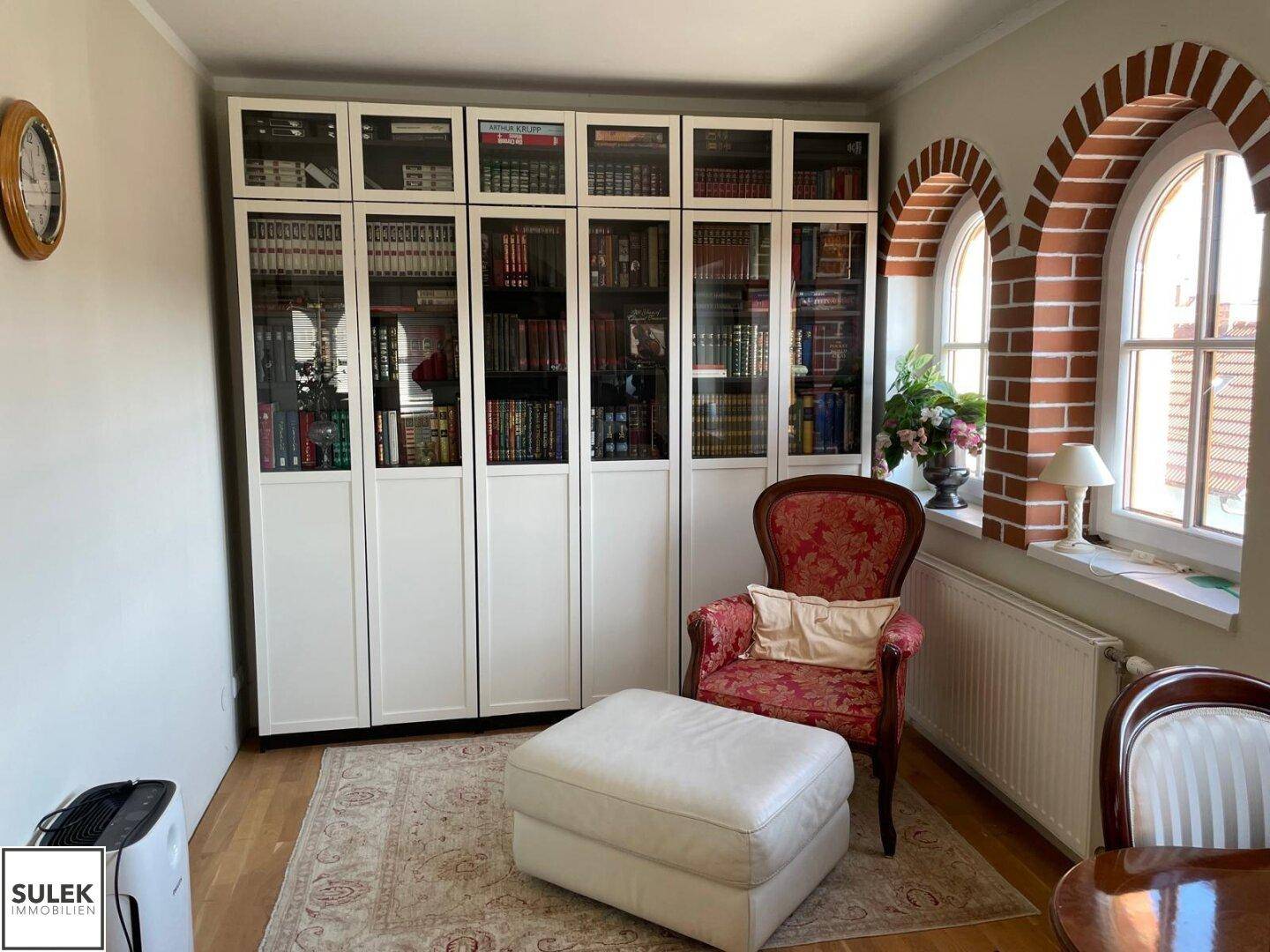 Wohnbereich mit Bücherregalen, bequemen Sesseln und einem Fenster mit dekorativem Ziegelbogen.