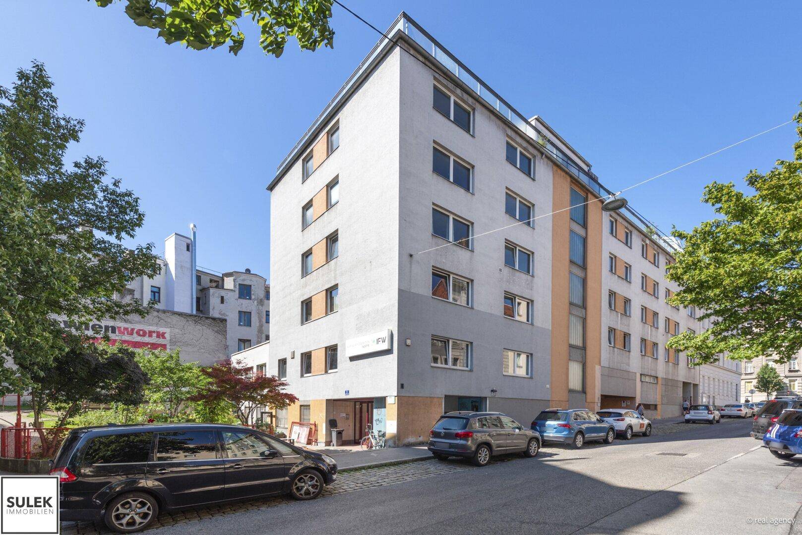 Mehrstöckiges Gebäude mit moderner Fassade und geparkten Autos an einer städtischen Straße.