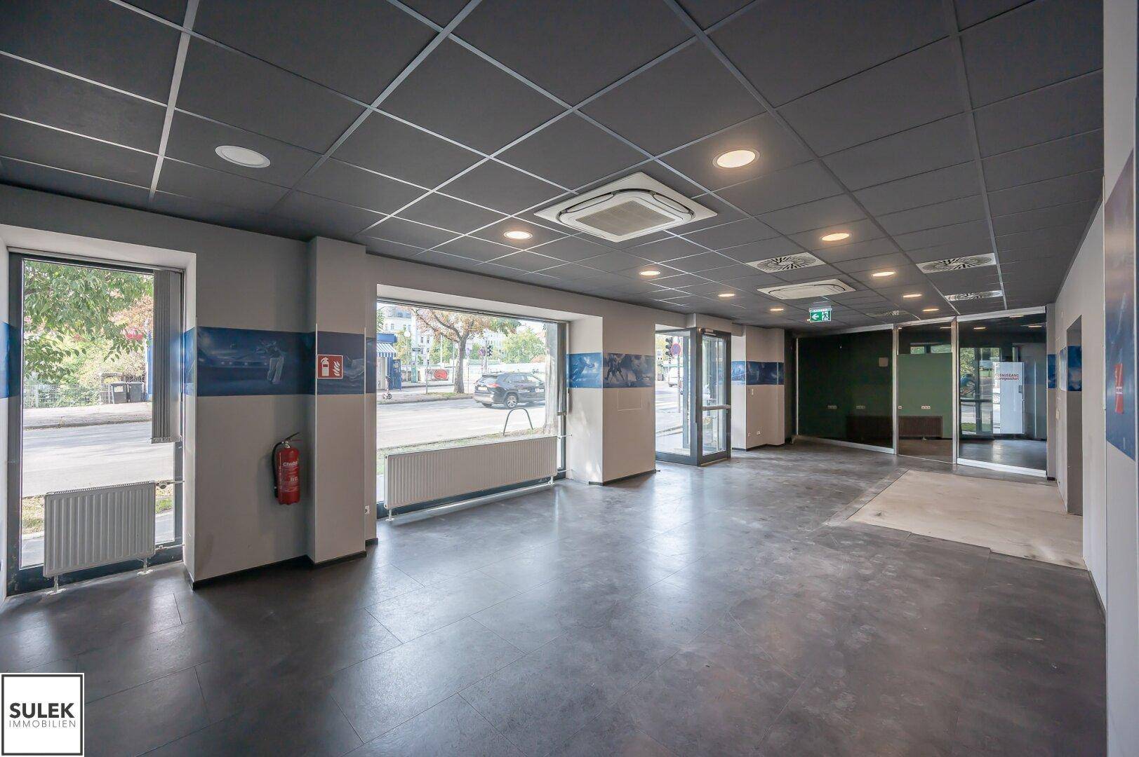Heller Gewerberaum mit großen Schaufenstern, dunklem Boden und abgehängter Decke mit Beleuchtung.
