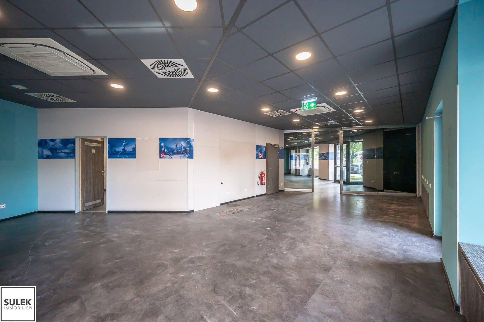 Weitläufiger Gewerberaum mit mehreren Türen, Glaswänden und einer abgehängten Decke.