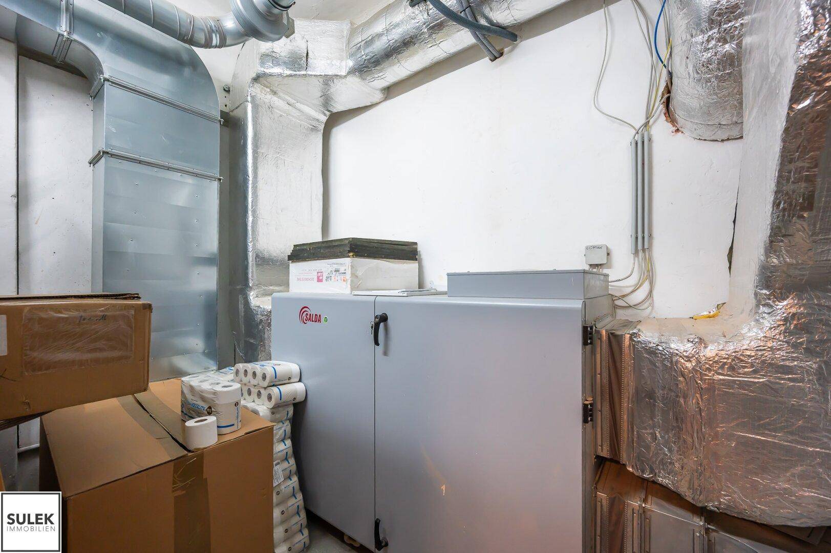 Technikraum im Keller mit Heizungsanlage, isolierten Rohren und Lagerkartons.