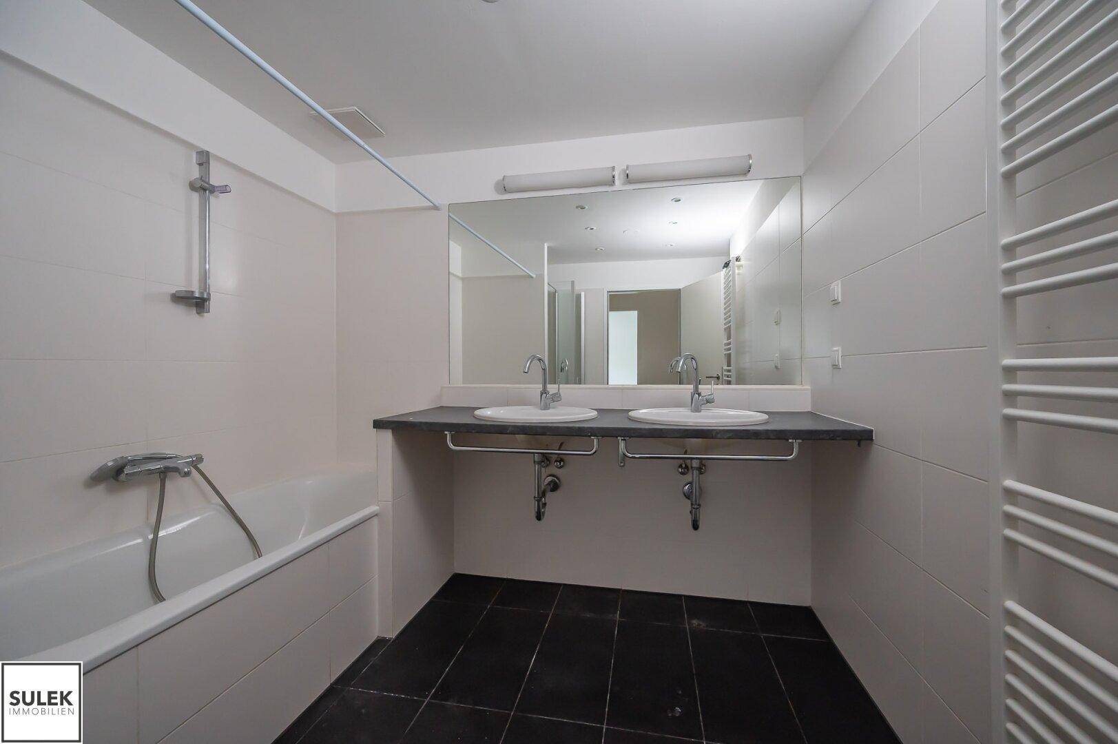 Geräumiges Badezimmer mit Badewanne, Doppelwaschbecken und weißen Fliesen an Wänden und Boden.