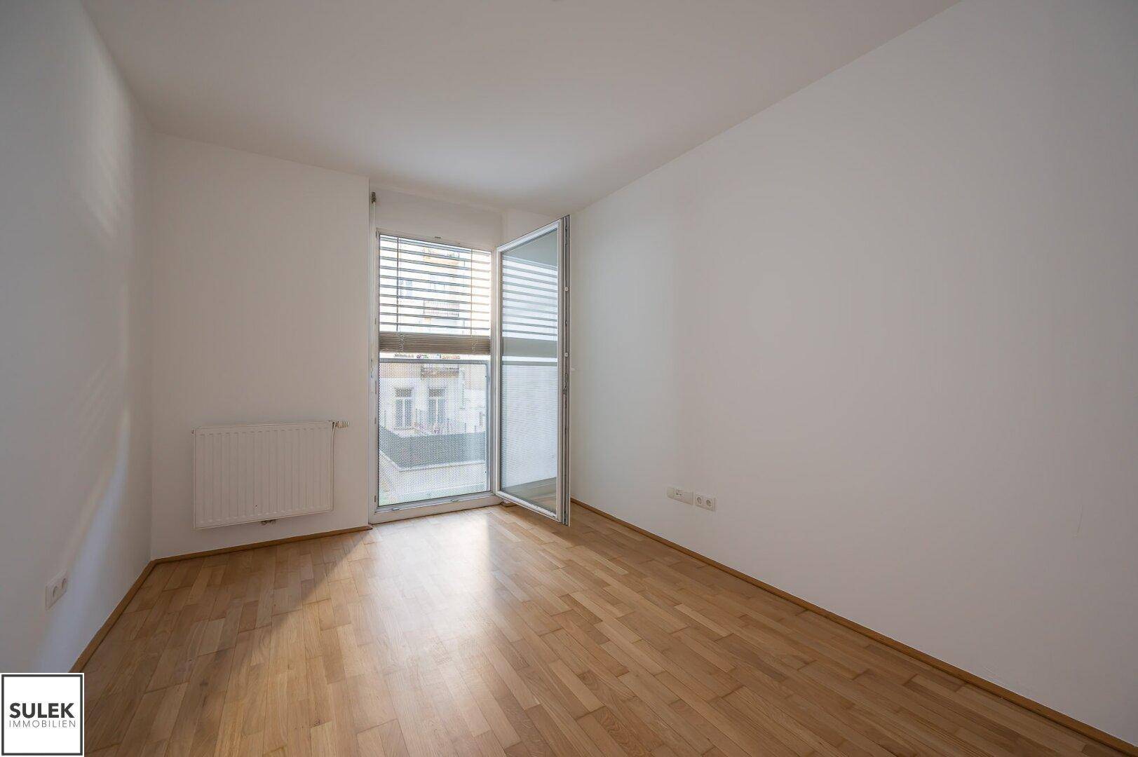 Helles Schlafzimmer mit Parkettboden, weißen Wänden und Zugang zu einem kleinen Balkon.