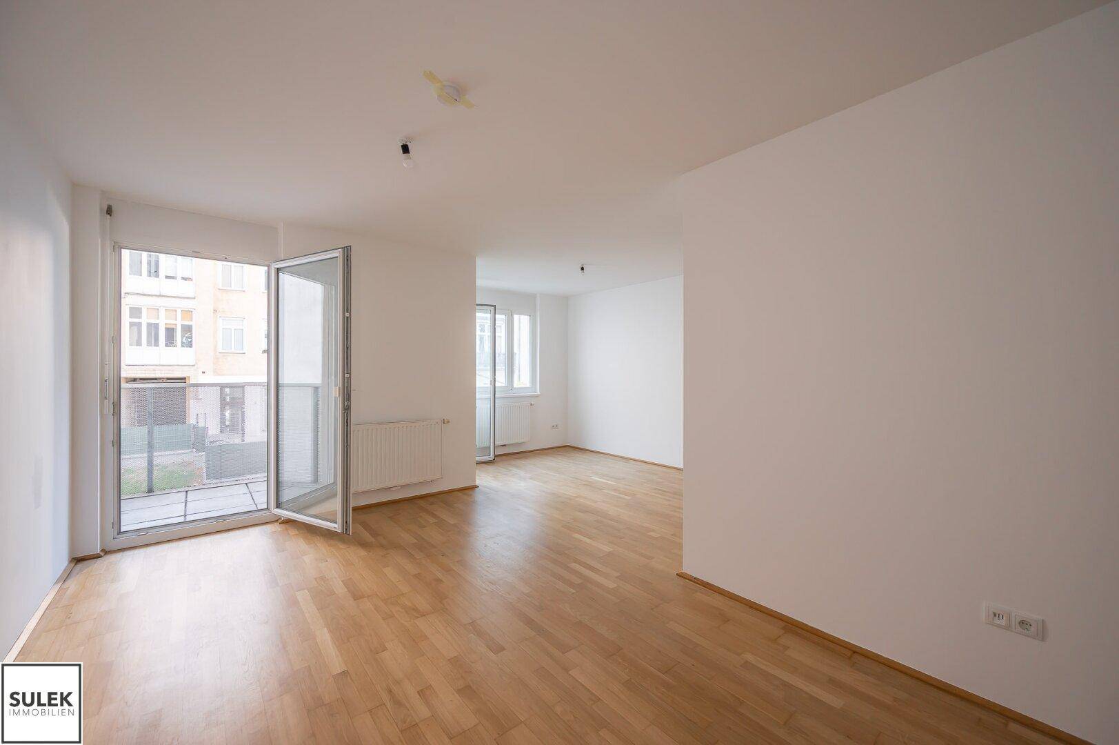 Heller, leerer Wohnraum mit Parkettboden, weißen Wänden und Zugang zu einem Balkon.