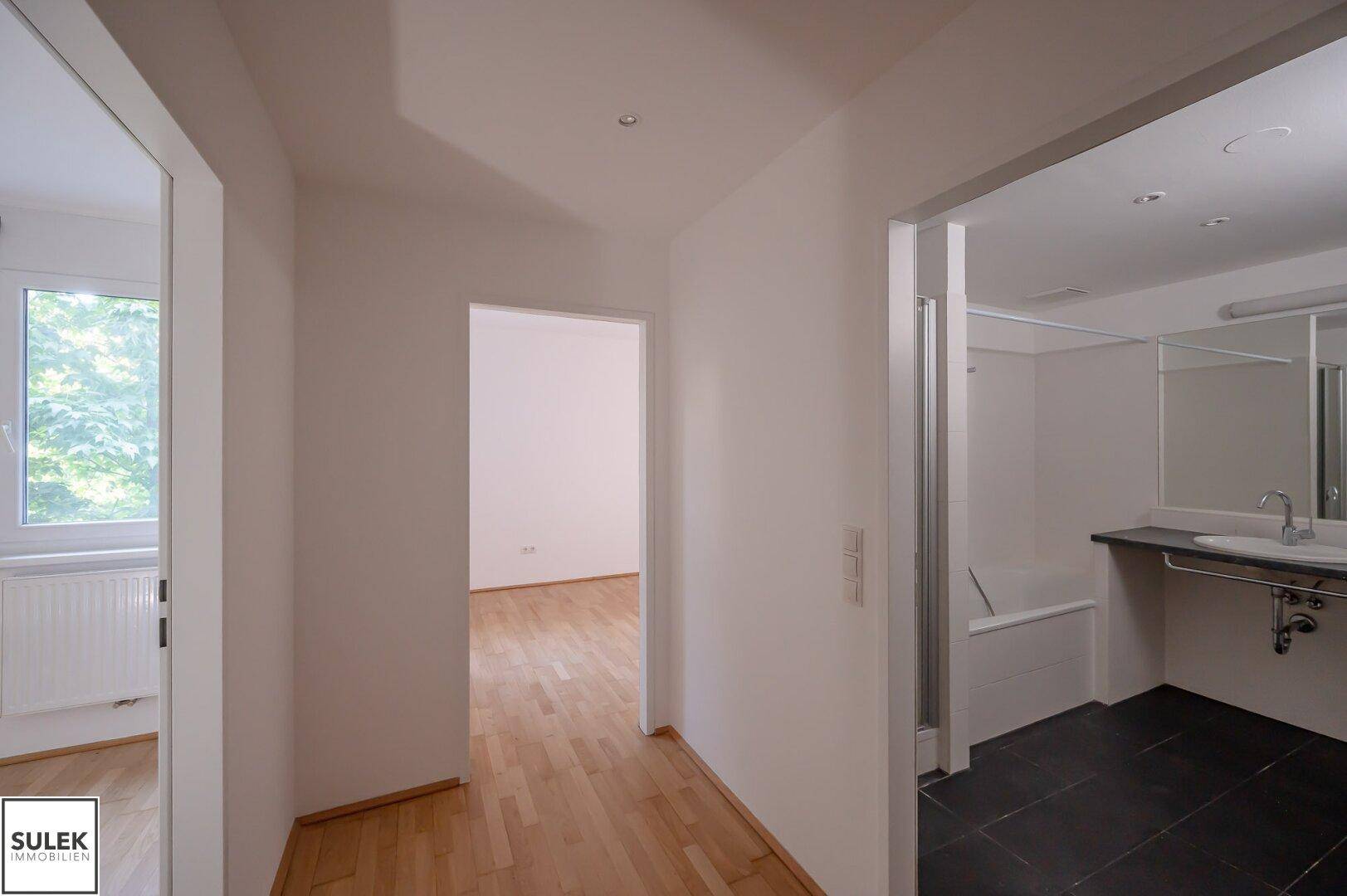 Flur mit Parkettboden, der zu einem Zimmer und einem Badezimmer mit Badewanne führt.