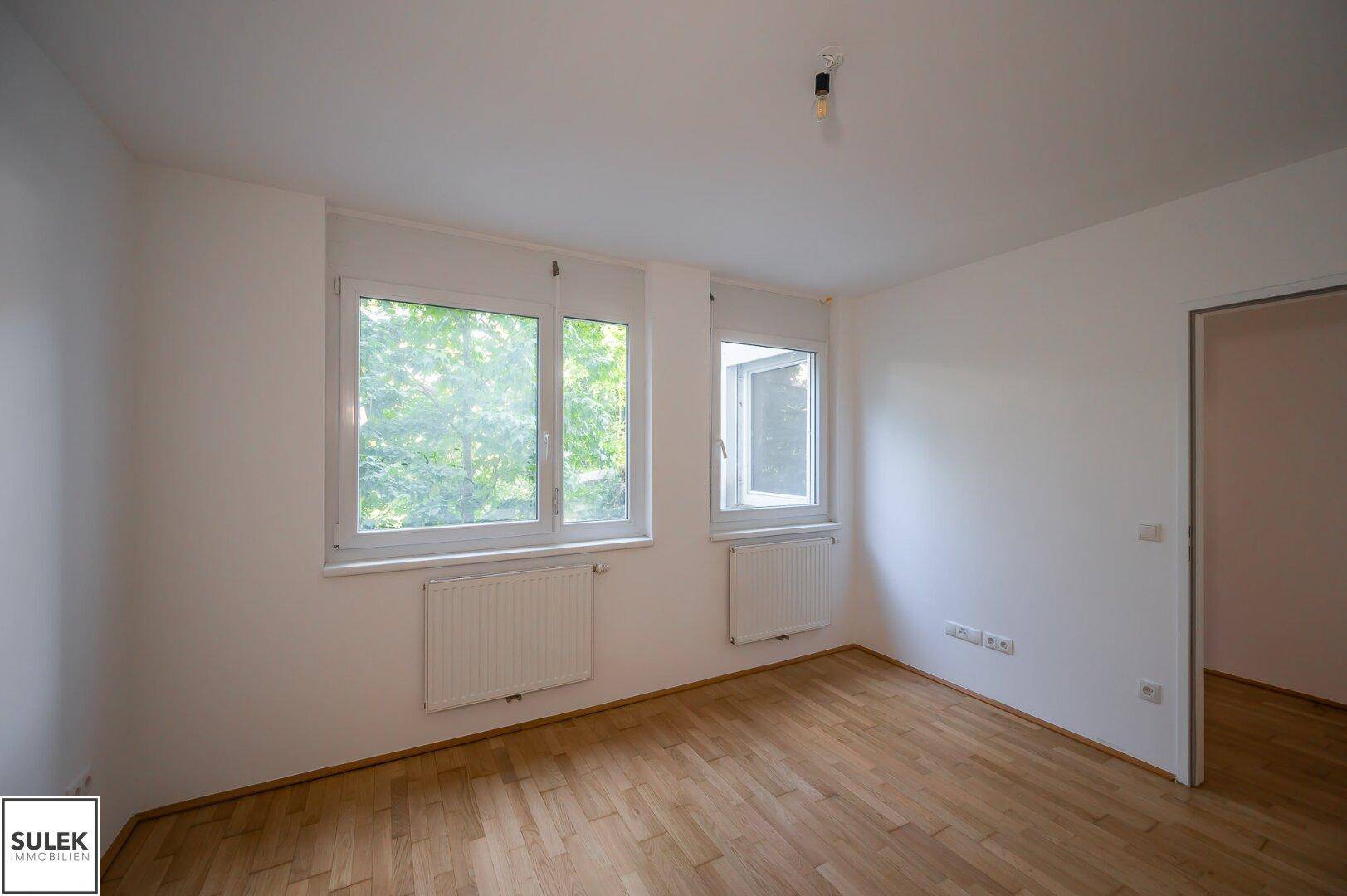 Helles Zimmer mit Parkettboden und zwei Fenstern, die einen Blick ins Grüne ermöglichen.