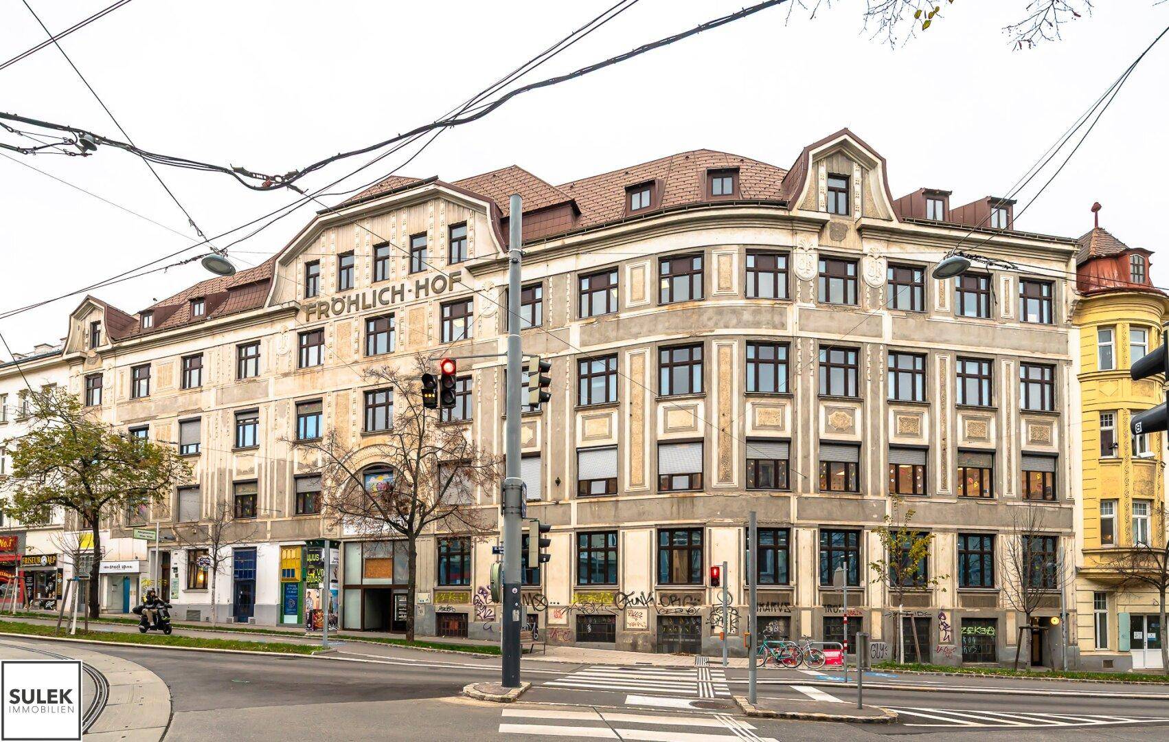 Ansicht des historischen Gebäudes "Fröhlich-Hof" von der Straßenseite mit Oberleitungen und Verkehr.