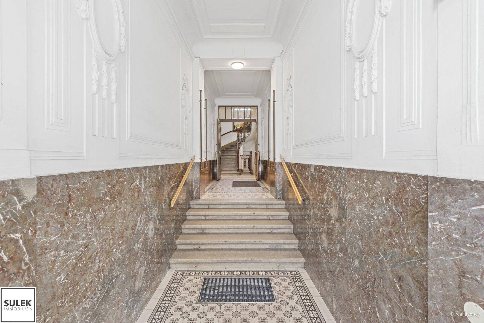 Elegante Eingangshalle mit Marmorverkleidung, gemustertem Fliesenboden und einer beeindruckenden Treppe im Hintergrund.