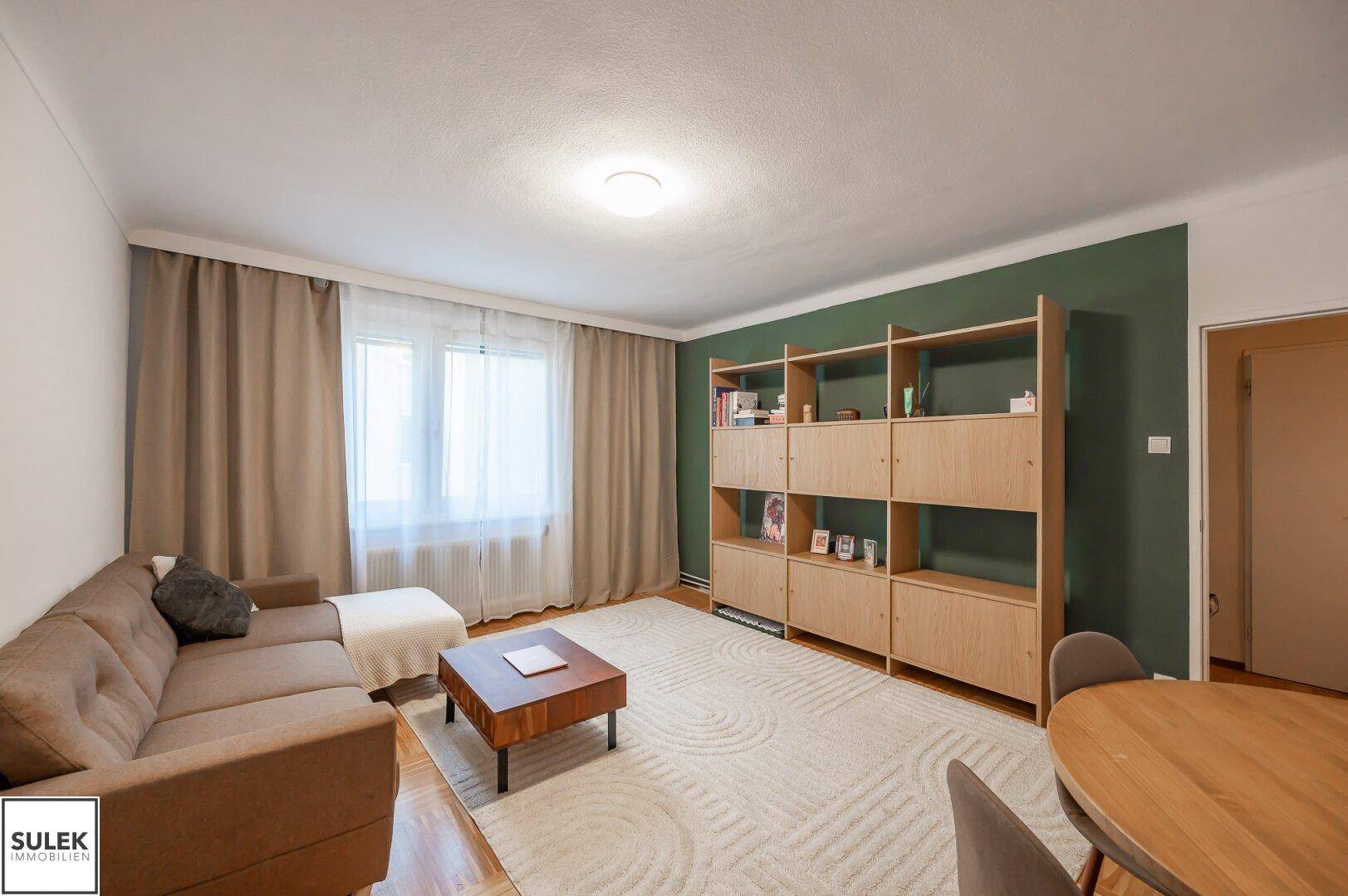 Heller Wohn- und Essbereich mit beigem Sofa, Holztisch und grüner Akzentwand.