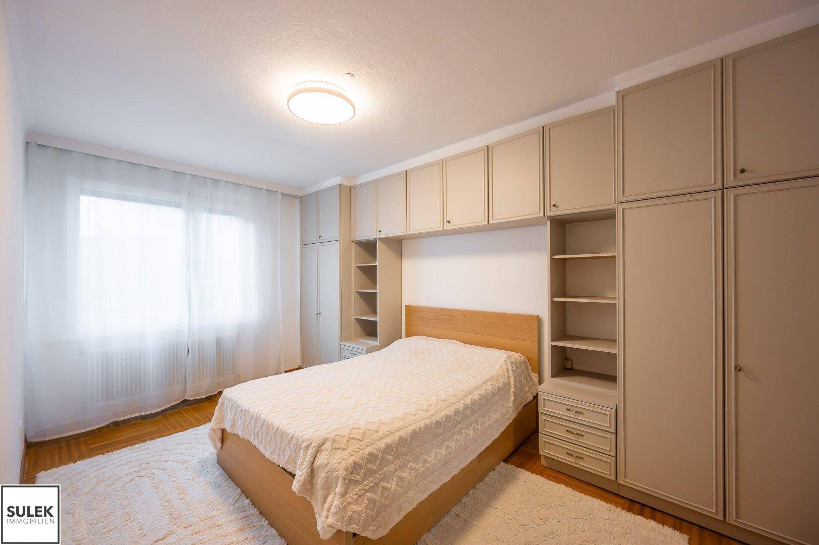 Helles Schlafzimmer mit großem Fenster, Vorhängen und praktischen Einbauschränken.
