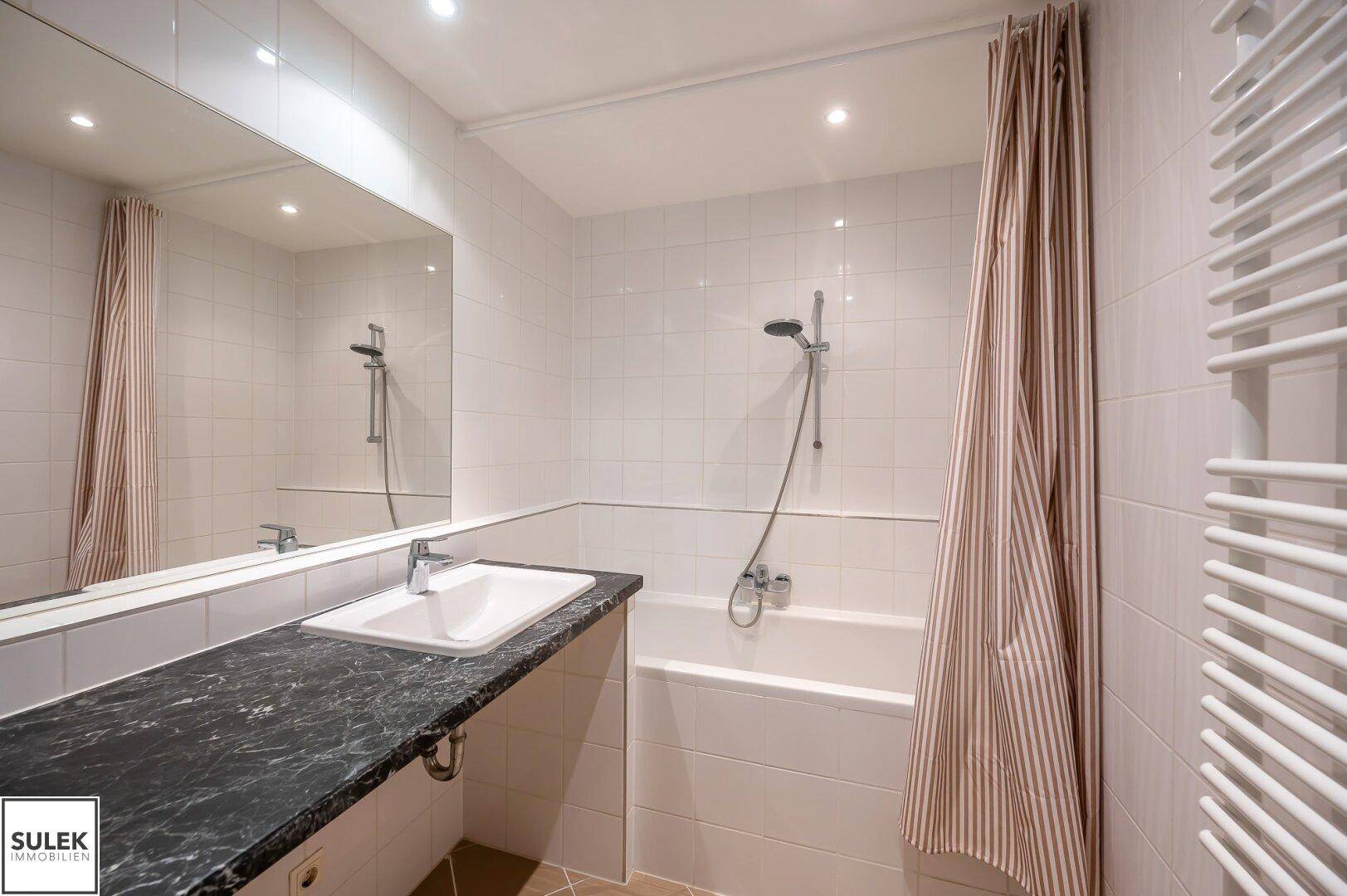 Modernes Badezimmer mit Badewanne, großem Spiegel, dunkler Arbeitsplatte und Handtuchheizkörper.