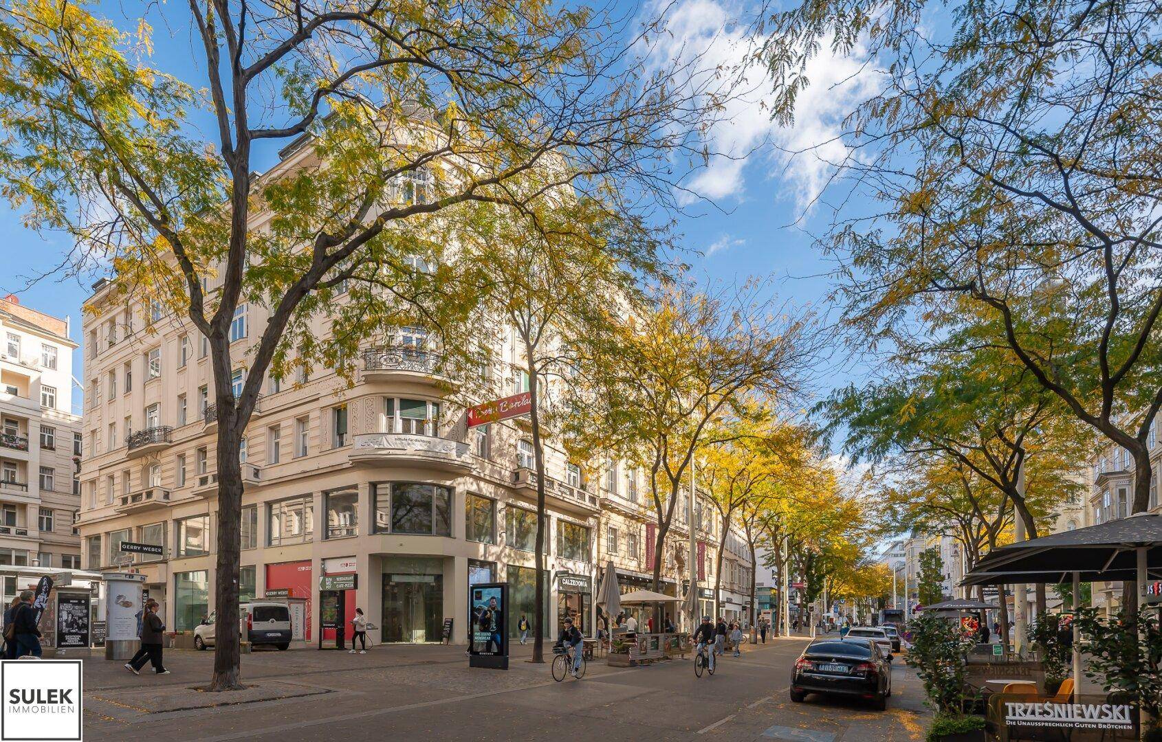 Historisches Gebäude an einer belebten Straße mit Bäumen und Geschäften, unter blauem Himmel.