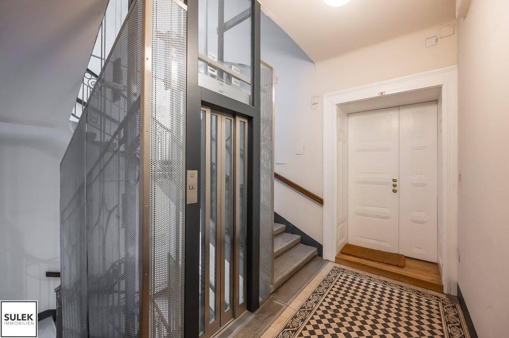 Moderner Aufzug aus Glas und Metall neben einer Treppe in einem historischen Gebäude mit gemustertem Boden.