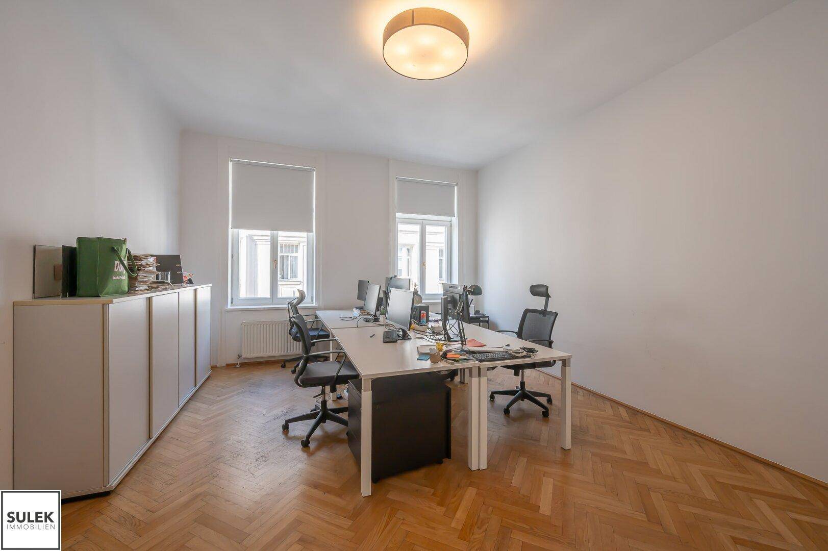 Großes Büro mit mehreren Arbeitsplätzen, weißen Schränken und Fischgrätparkett, gut beleuchtet durch Fenster.