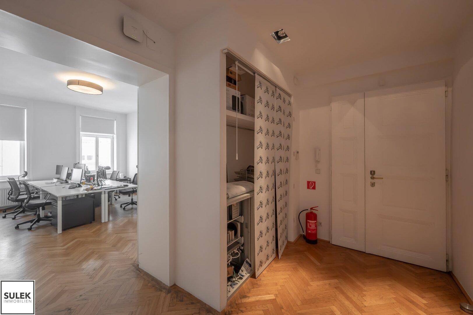 Flur mit Fischgrätparkett, Einbauschrank und Zugang zu einem Büroraum, ausgestattet mit Feuerlöscher.