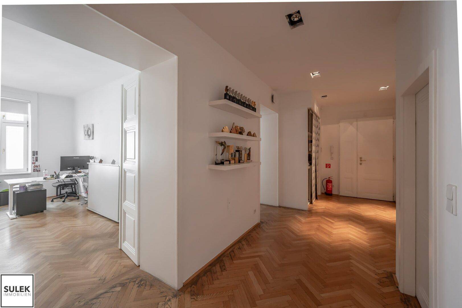 Geräumiger Flur mit Fischgrätparkett, weißen Türen und Blick in einen angrenzenden Büroraum.