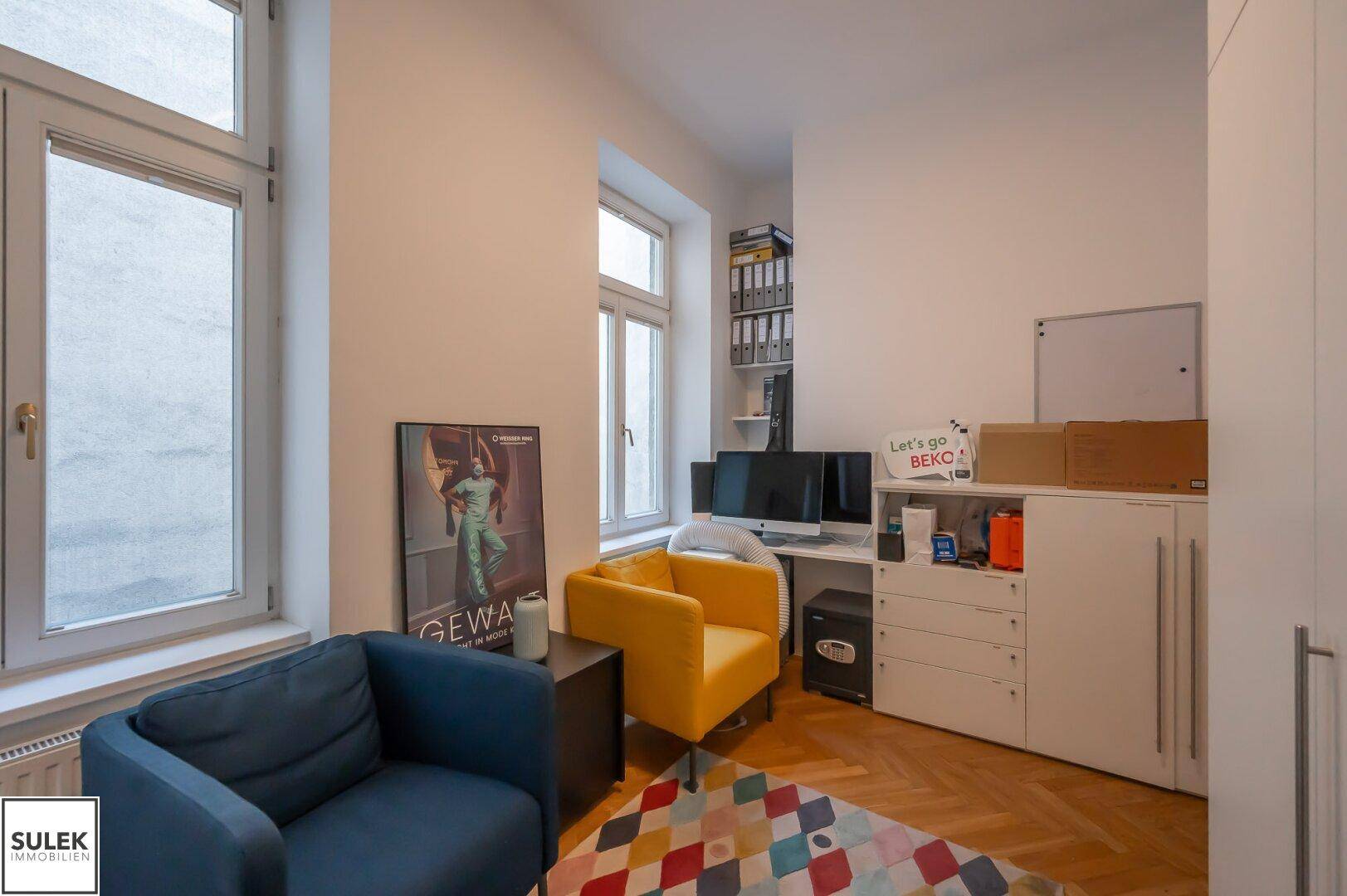Büroraum mit Einbauschränken, Fischgrätparkett und zwei Fenstern, ausgestattet mit einem Schreibtisch und Stühlen.