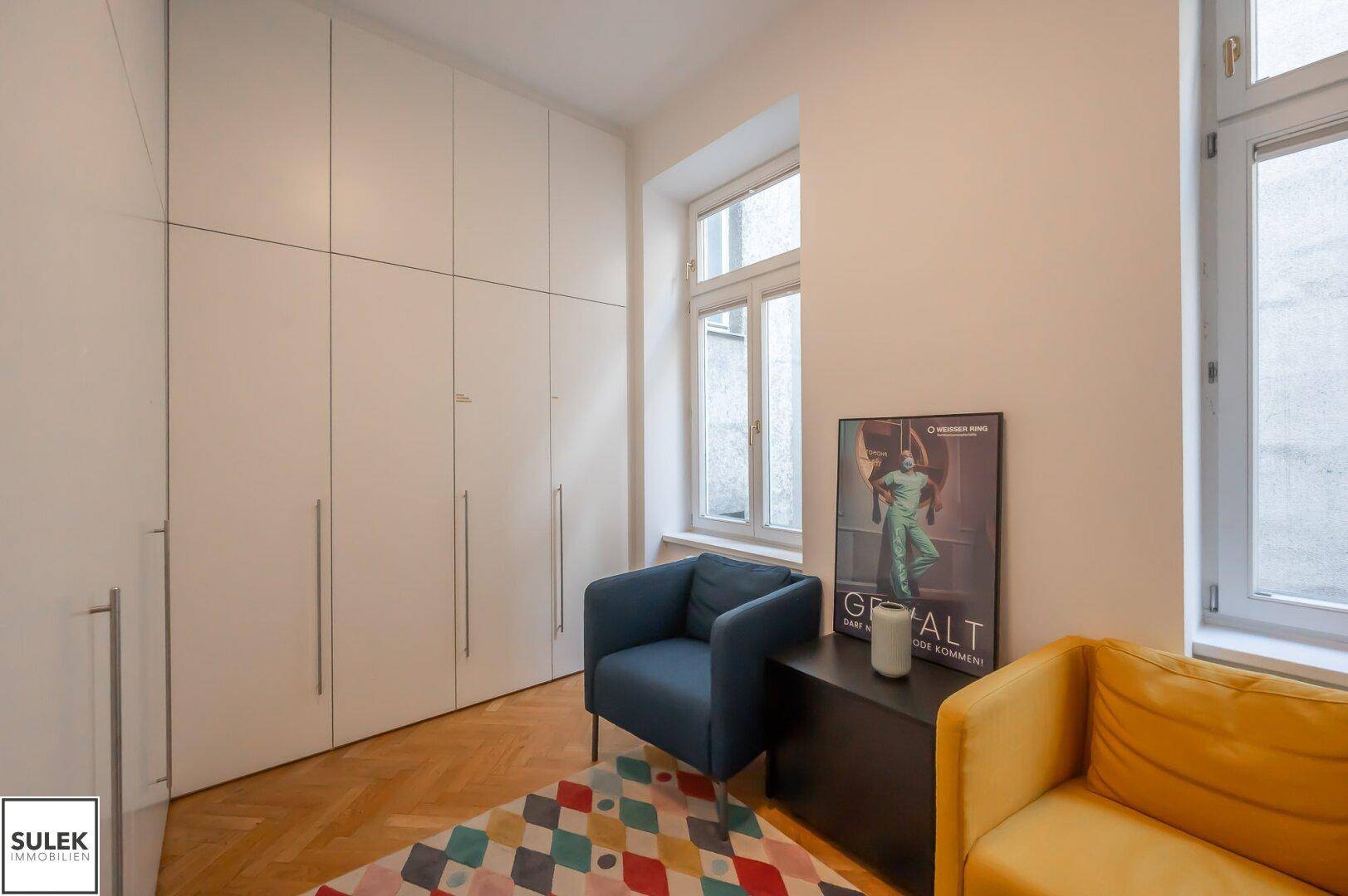 Schlafzimmer mit Einbauschränken, Fischgrätparkett und zwei Fenstern, dekoriert mit einem bunten Teppich.