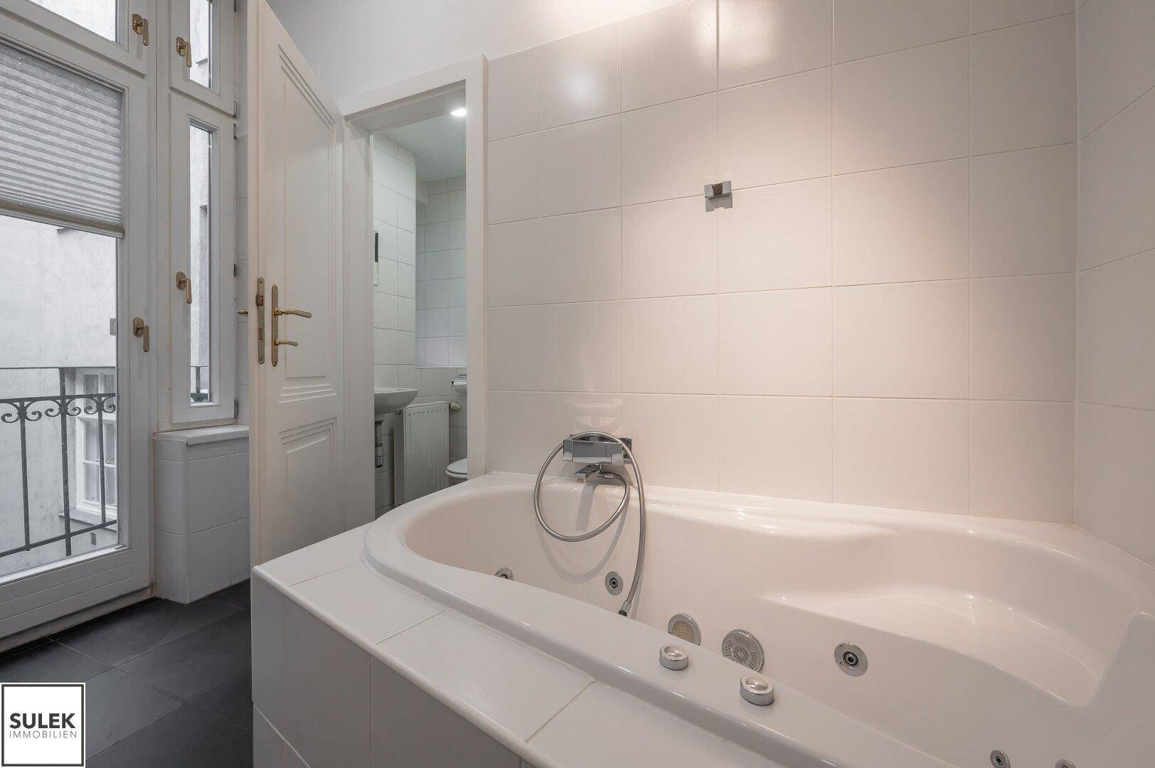 Badezimmer mit Whirlpool-Badewanne und weißen Fliesen, mit Zugang zu einem kleinen Balkon.