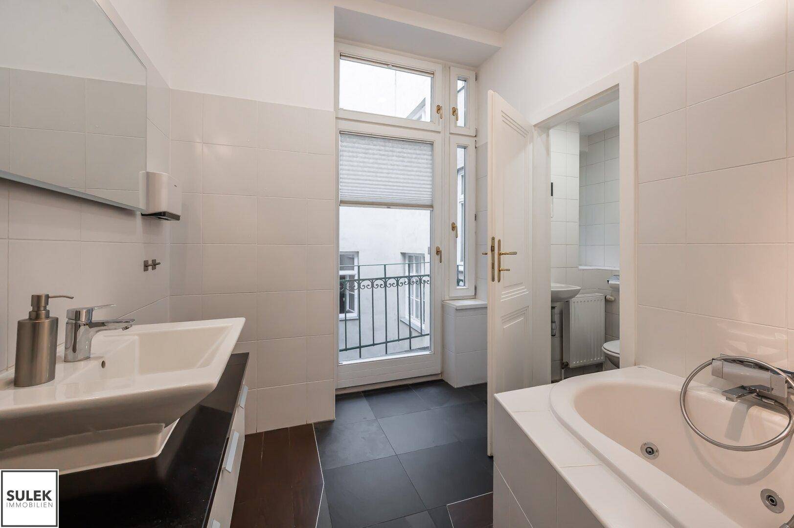 Badezimmer mit Badewanne, Waschbecken und Zugang zu einem kleinen Balkon, hell gefliest.