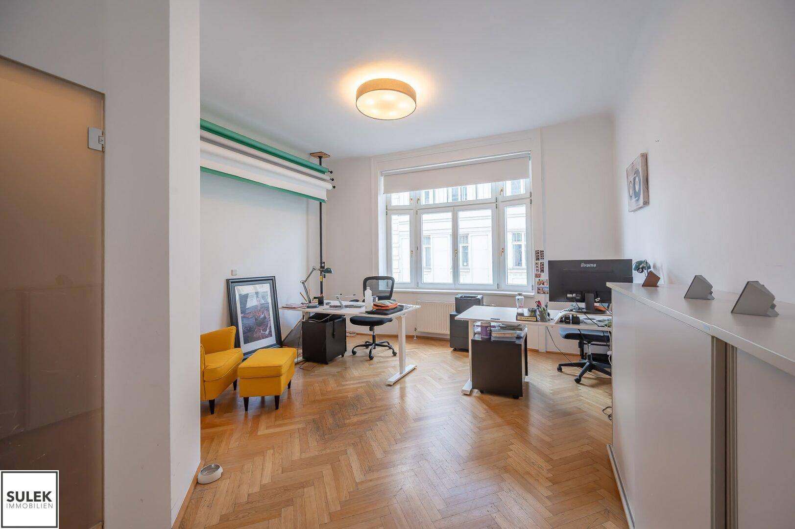 Helles Büro mit Fischgrätparkett, großen Fenstern und mehreren Arbeitsplätzen, ausgestattet mit Büromöbeln.