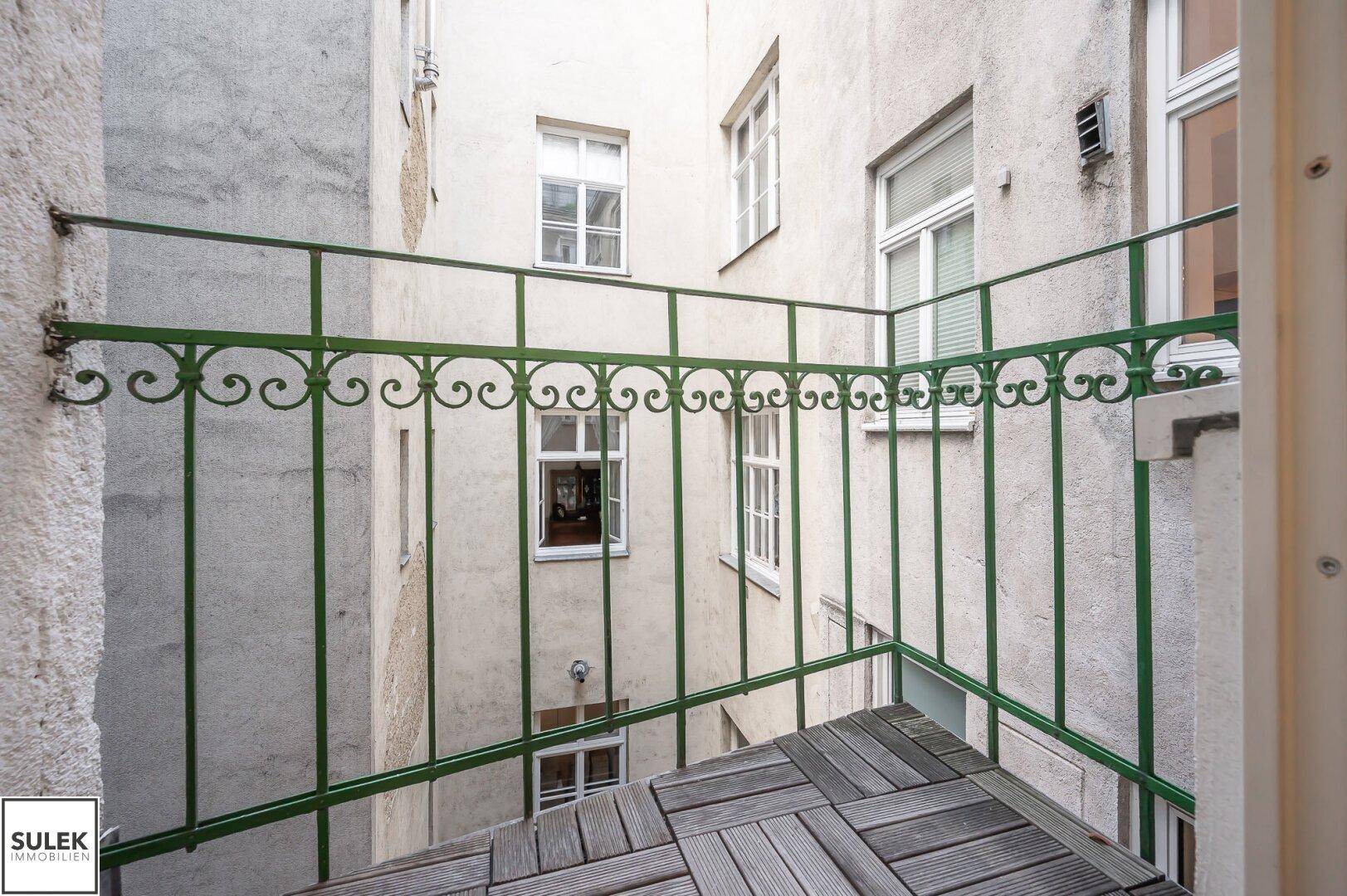 Kleiner Balkon mit Holzboden und schmiedeeisernem Geländer, mit Blick auf einen Innenhof.