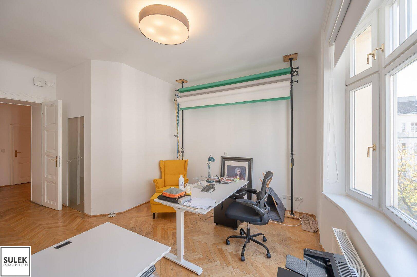 Büroraum mit Fischgrätparkett, großen Fenstern und einem Arbeitsplatz, ergänzt durch eine gelbe Sitzgelegenheit.