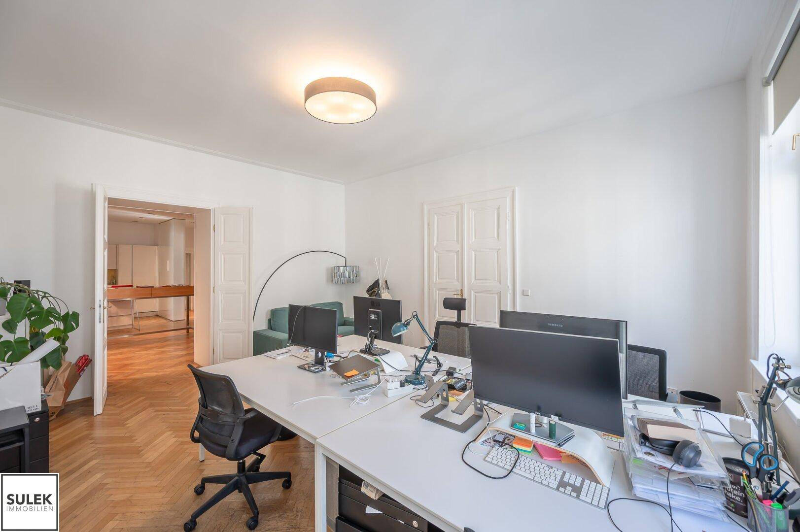 Büroraum mit mehreren Arbeitsplätzen, Fischgrätparkett und Blick in einen angrenzenden Bereich.