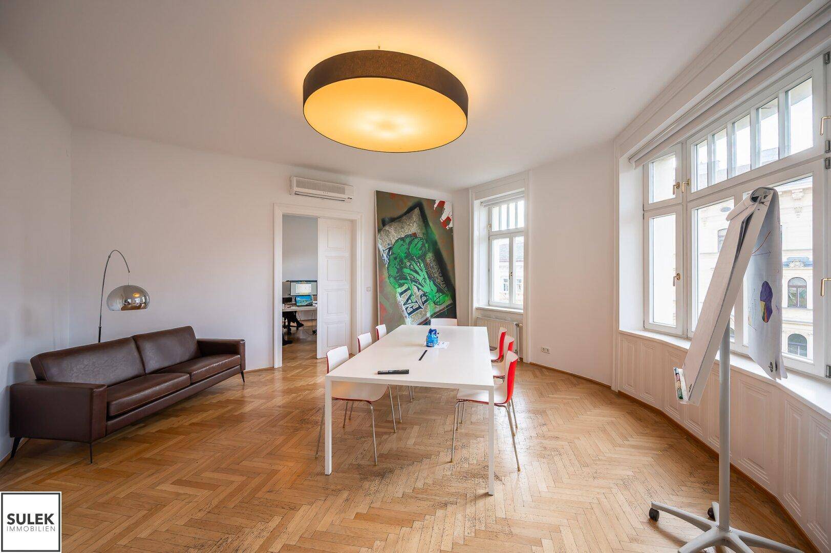Heller Wohn- oder Besprechungsraum mit Sofa, Esstisch und großen Fenstern, ausgestattet mit Fischgrätparkett.