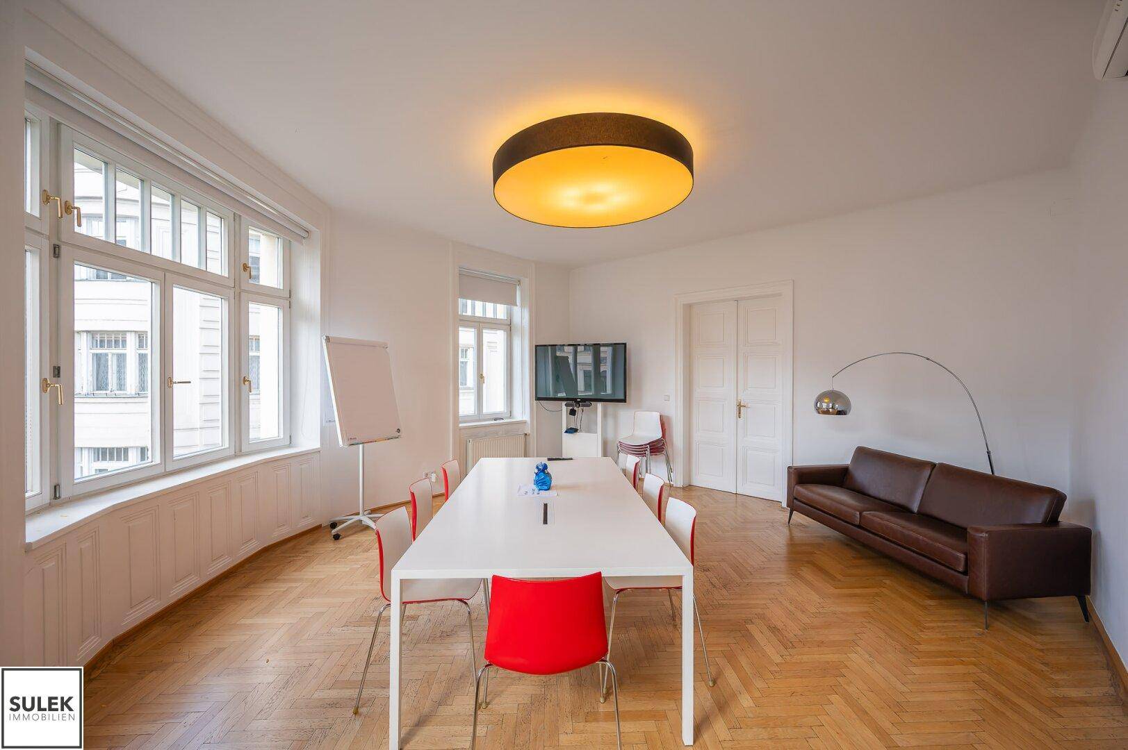 Heller Büroraum mit Konferenztisch, Ledersofa und Fischgrätparkett, ideal für Besprechungen und Pausen.
