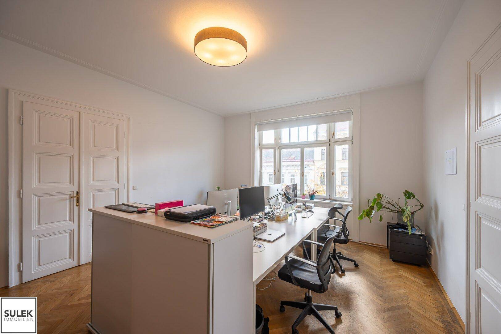 Helles Büro mit mehreren Arbeitsplätzen, großen Fenstern und klassischem Fischgrätparkettboden.
