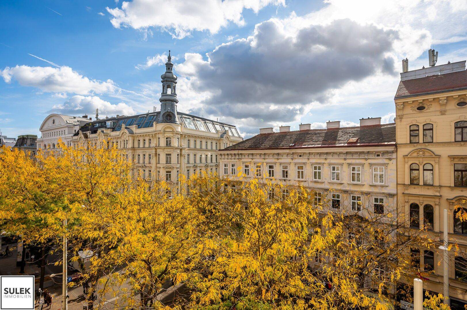 Historisches Gebäude mit markantem Turm und Herbstbäumen unter blauem Himmel.
