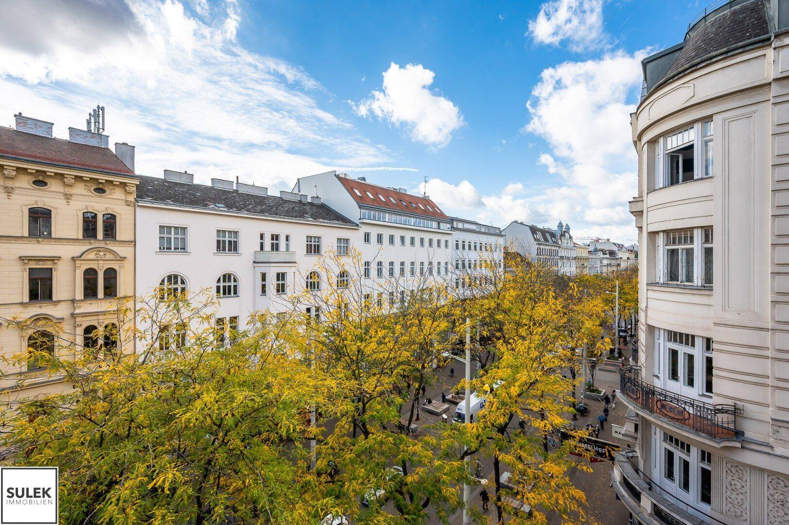 Panoramablick auf eine belebte Straße mit herbstlichen Bäumen und historischen Gebäuden unter blauem Himmel.