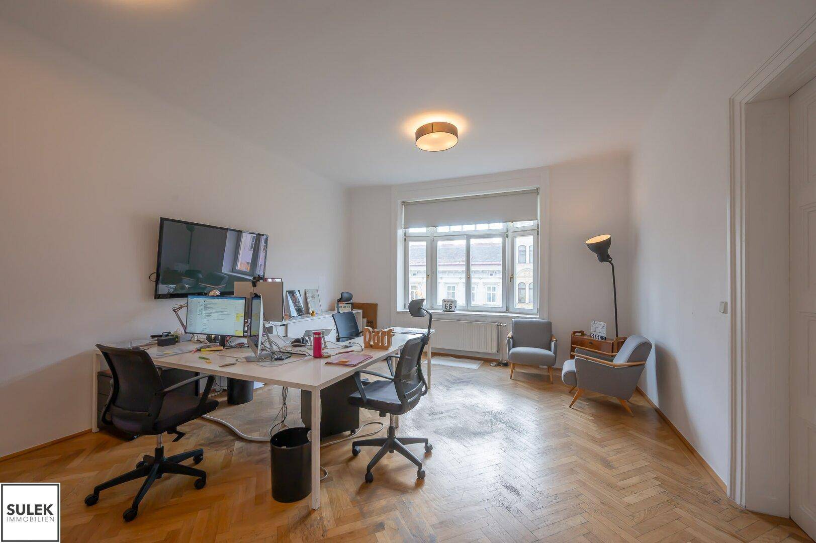 Funktionales Büro mit Arbeitsplätzen, Bildschirmen und bequemen Sesseln, beleuchtet durch große Fenster.
