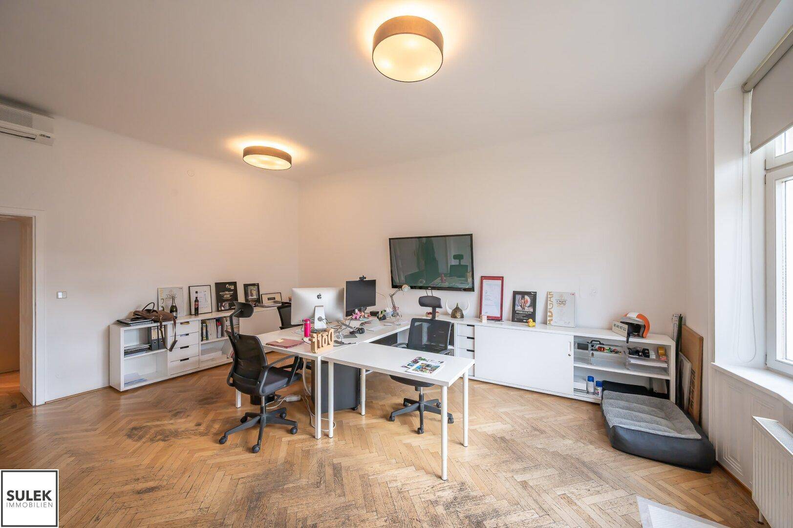 Großzügiges Büro mit mehreren Arbeitsbereichen, weißen Regalen und klassischem Fischgrätparkettboden.