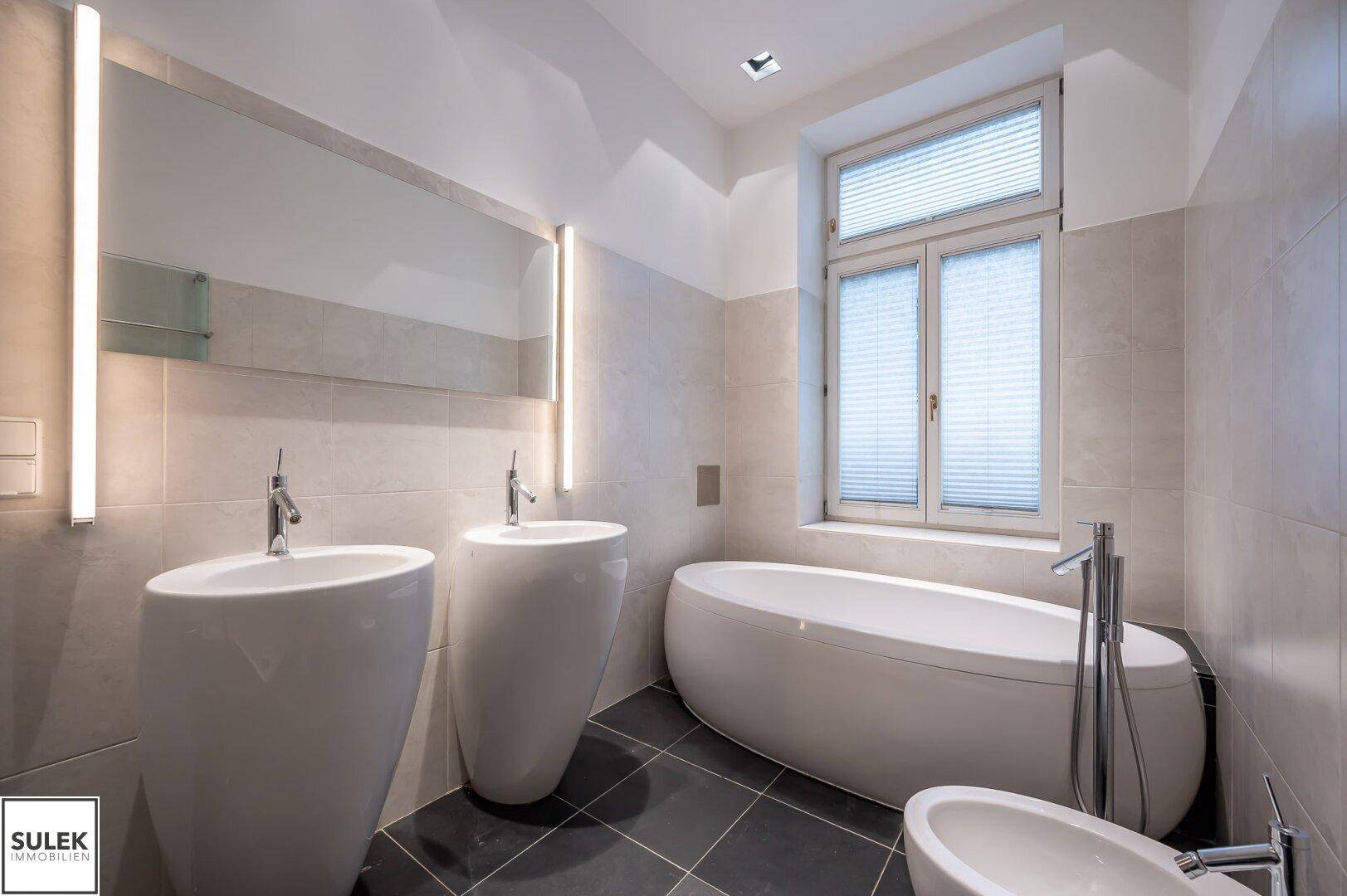 Modernes Badezimmer mit freistehender Badewanne, zwei Waschbecken und großem Spiegel, gut beleuchtet.