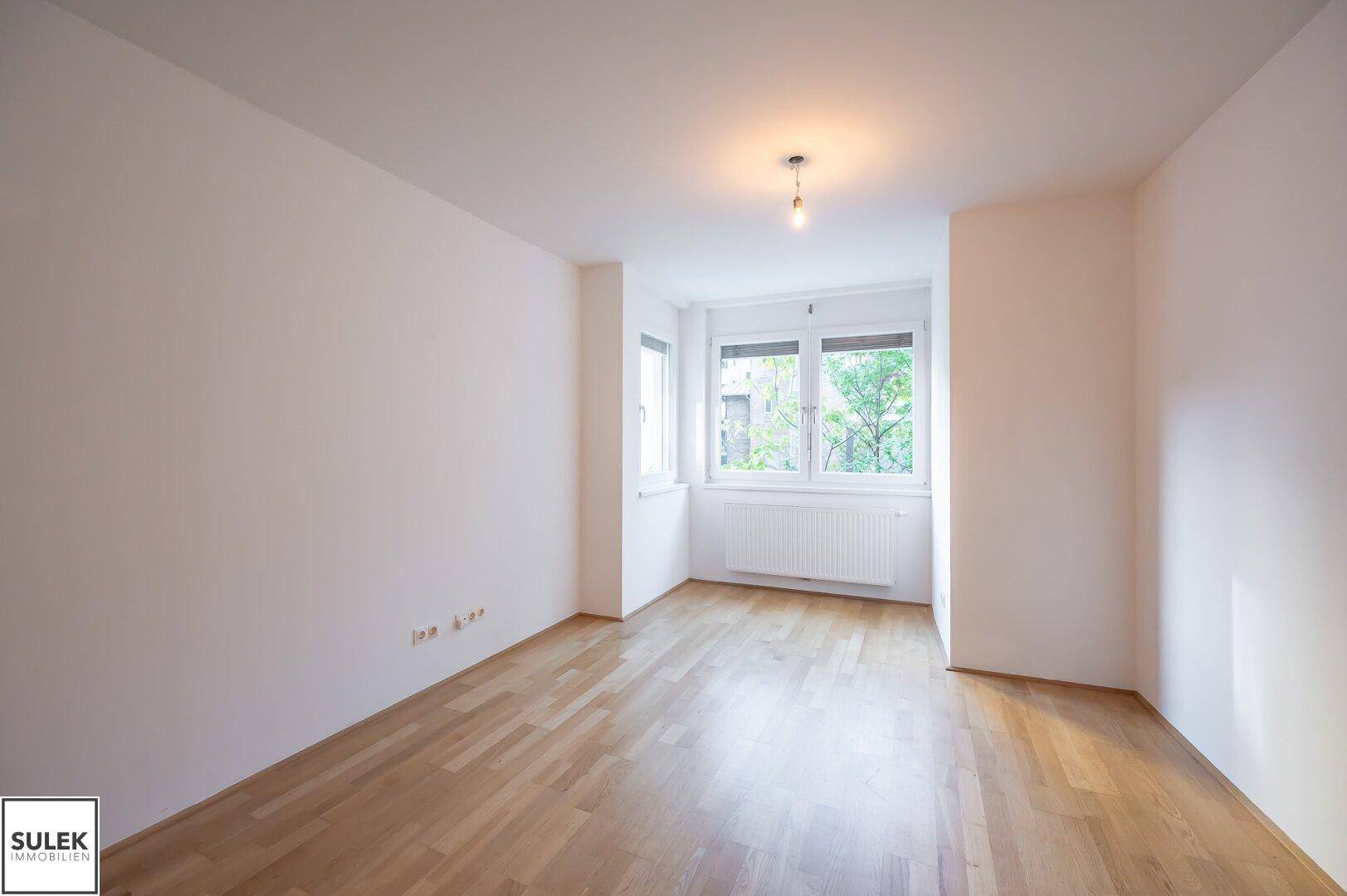 Leeres, helles Zimmer mit Holzboden und großen Fenstern, bereit zur individuellen Gestaltung.