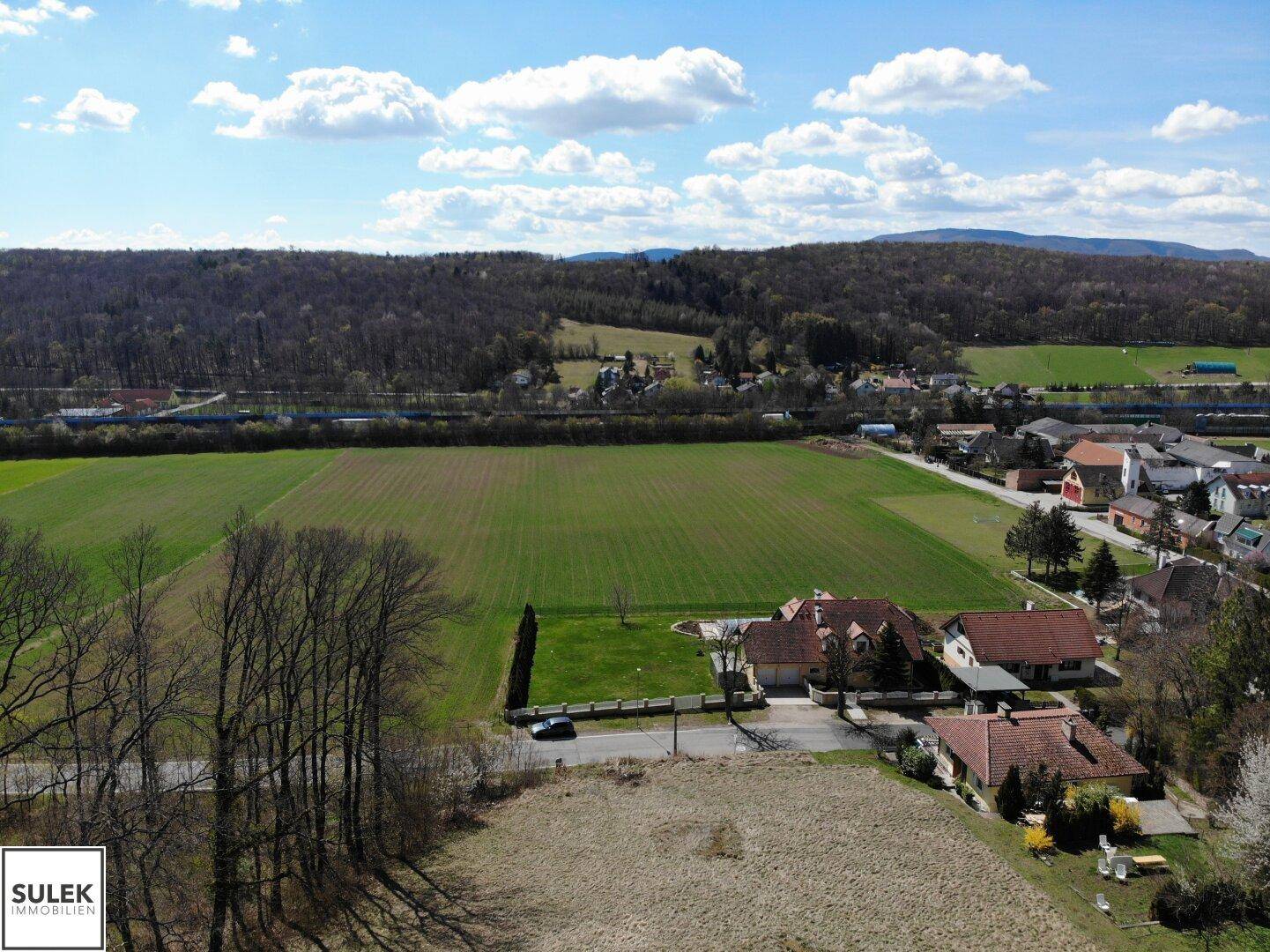 Panoramablick über ein weites Grundstück mit angrenzendem Dorf, grünen Feldern und bewaldeten Hügeln im Hintergrund.