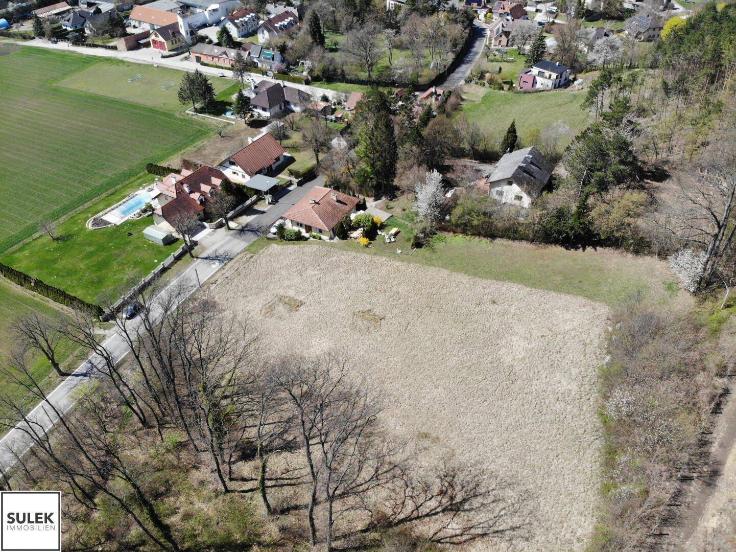 Luftaufnahme eines Baugrundstücks mit Blick auf benachbarte Häuser, darunter eines mit Pool, und weite Felder.
