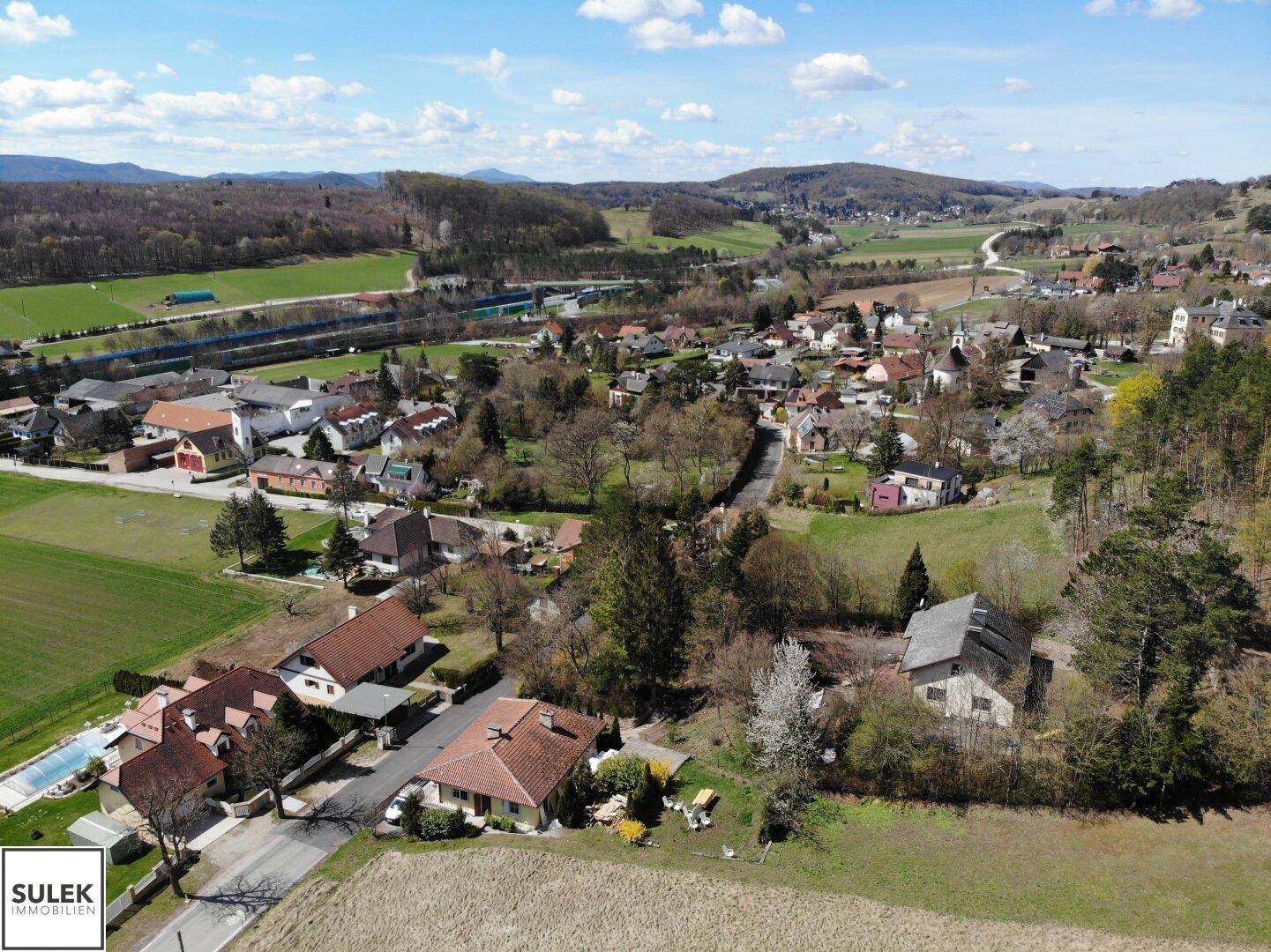 Ausgedehnte Luftaufnahme eines Dorfes mit umliegenden Feldern und Wäldern unter einem strahlend blauen Himmel.